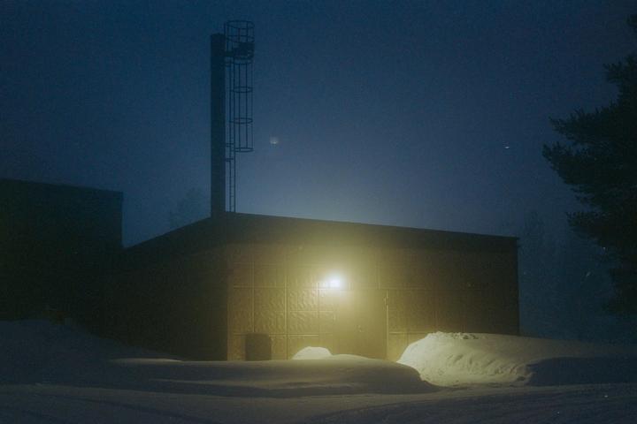A building with a tall chimney and a bright light on its facade stands in a snowy landscape at night.