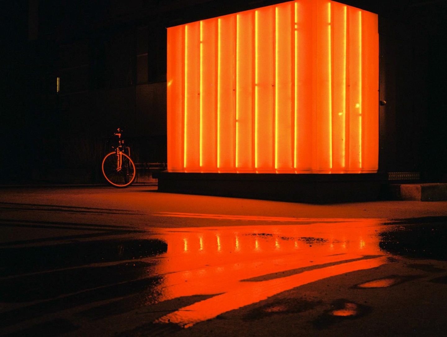 A bicycle is parked next to a large orange neon light box, reflecting on the wet ground at night.