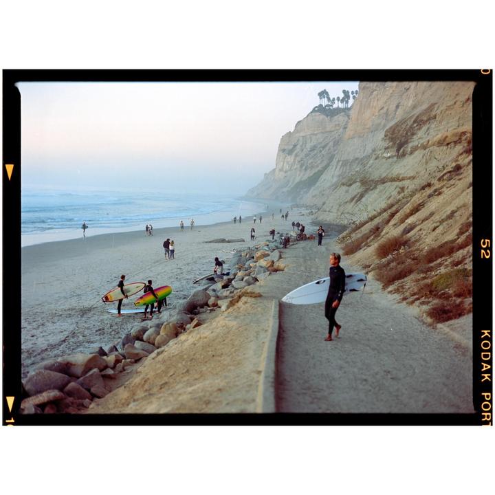 A surfer walks along a path next to a beach with cliffs in the background.