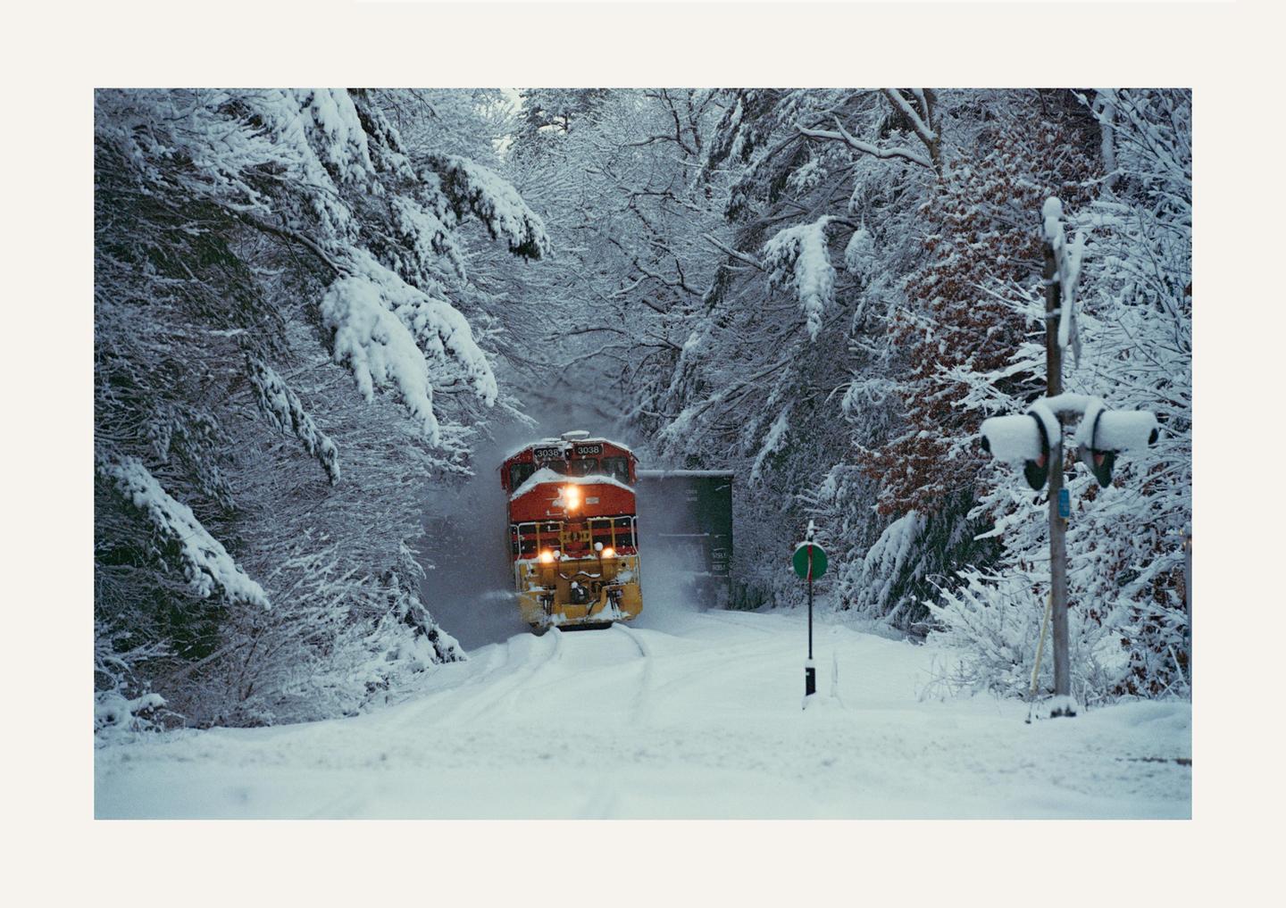 A red and yellow train emerges from a snowy forest, with snow-covered trees lining the tracks.