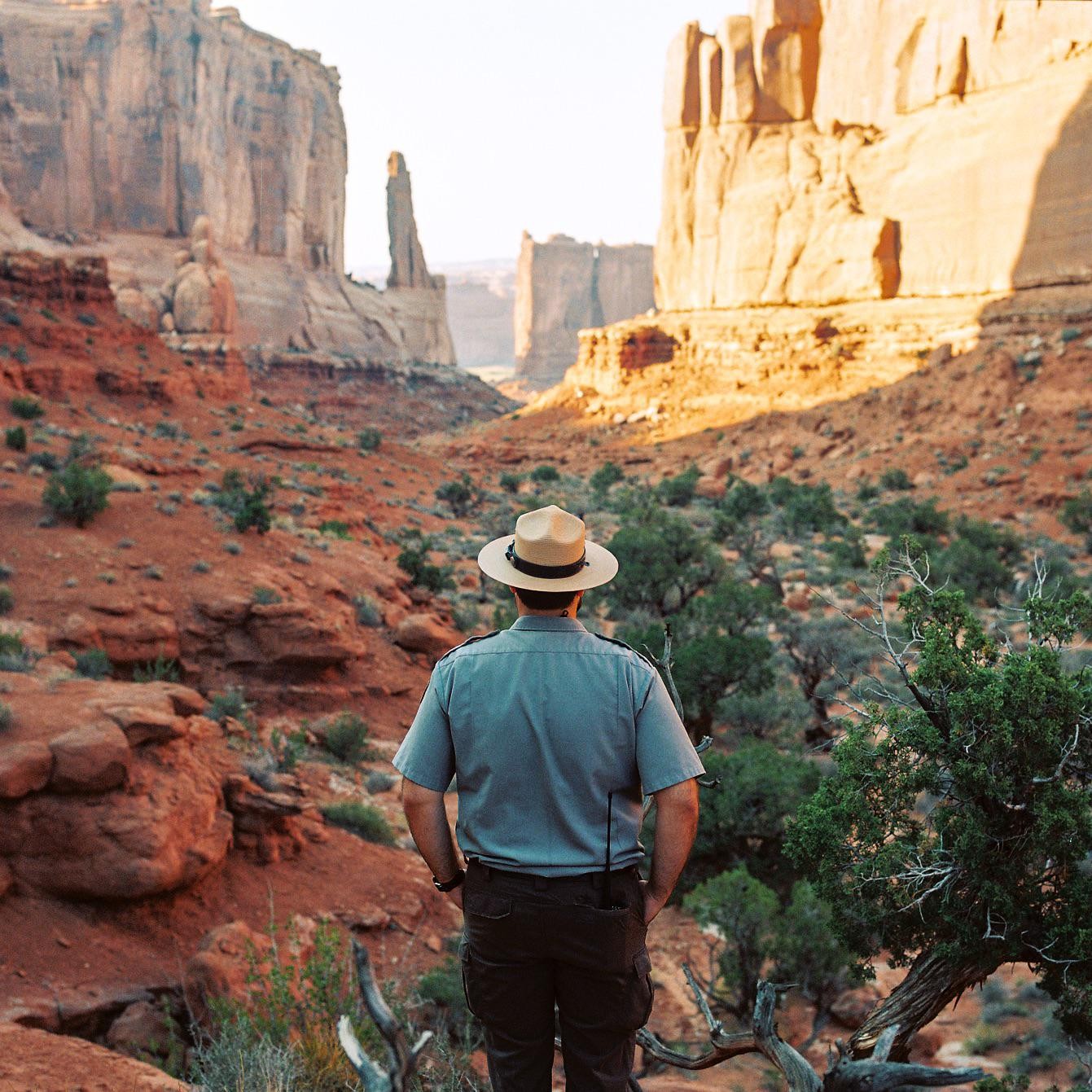 A man in a ranger uniform and hat stands with his back to the camera, looking out at a desert landscape with large rock formations.