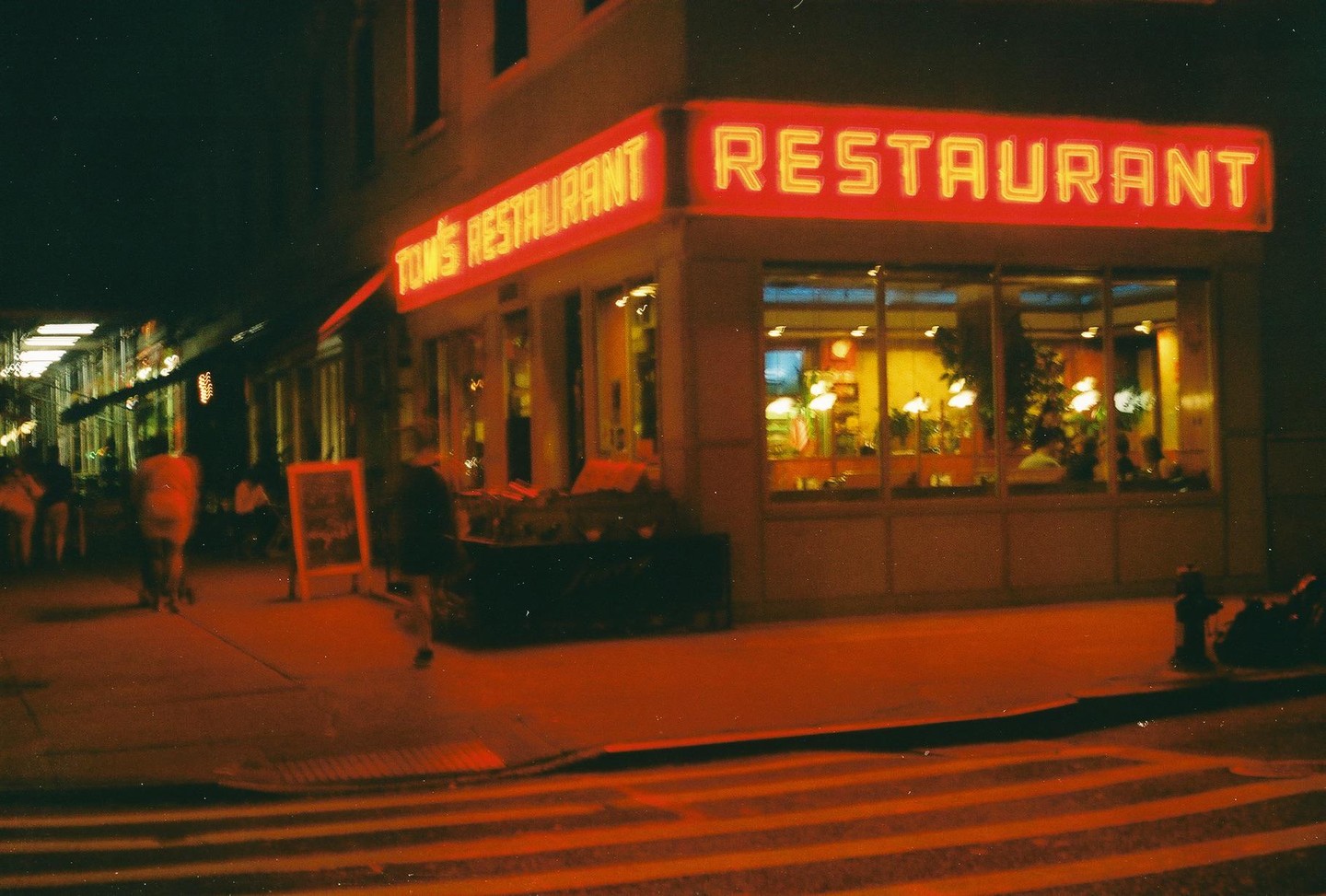 A restaurant with neon signs is visible at night on a street corner with people walking by.