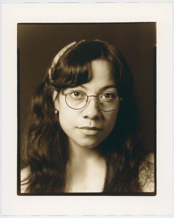 A woman with glasses and long, dark hair looks directly at the viewer.