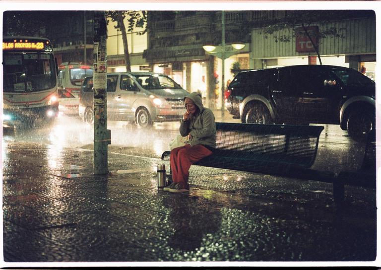 A person in a hooded sweatshirt sits on a bench in the rain at night, with cars and a bus passing by on a wet street.