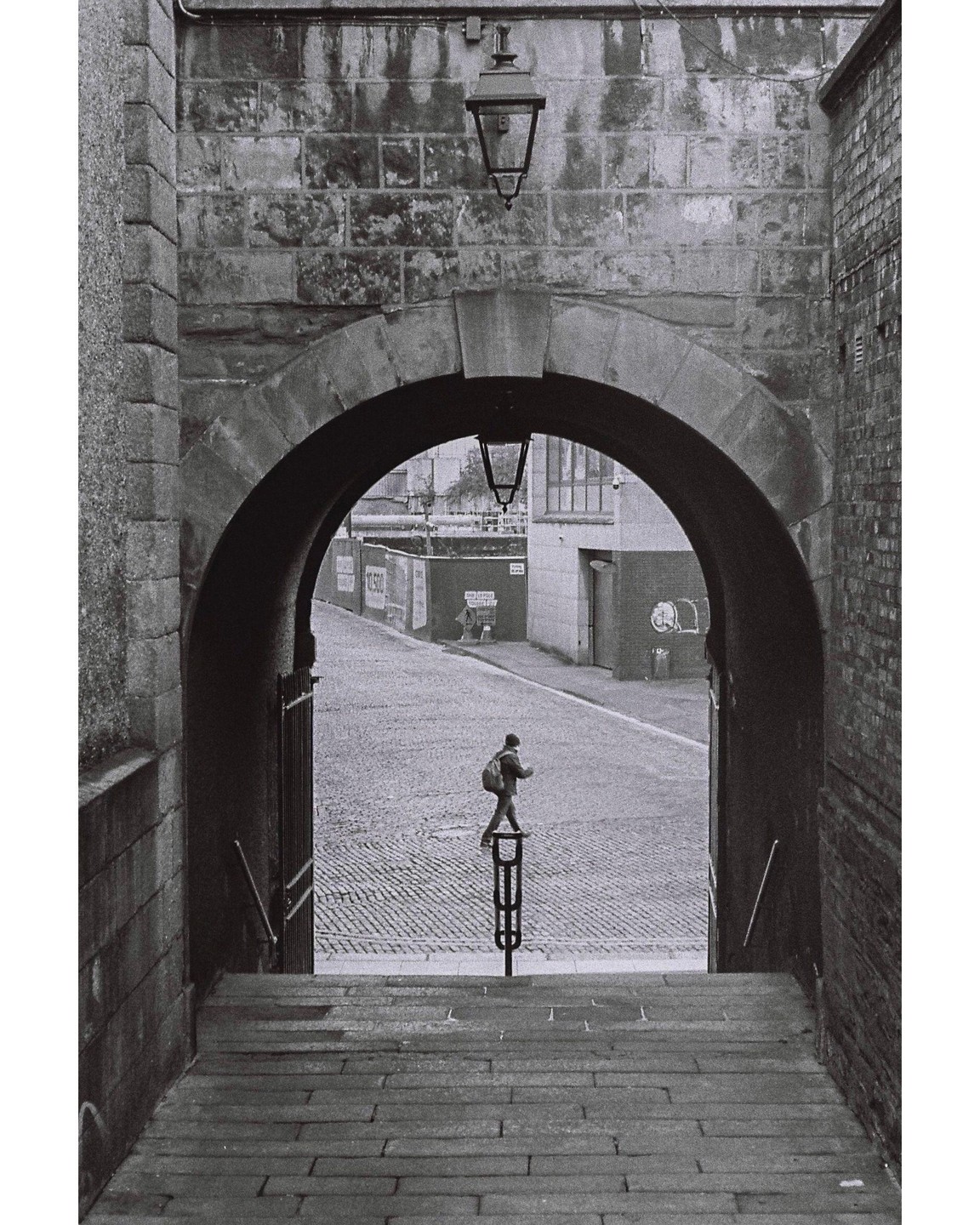 A person with a backpack walks on a cobblestone street through a stone archway.