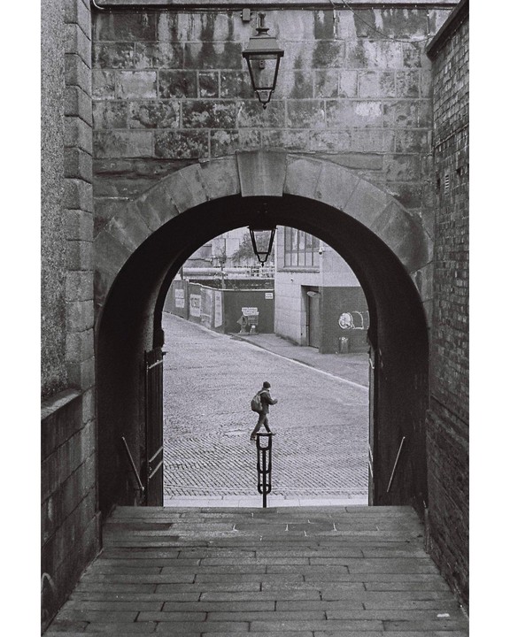 A person with a backpack walks on a cobblestone street through a stone archway.