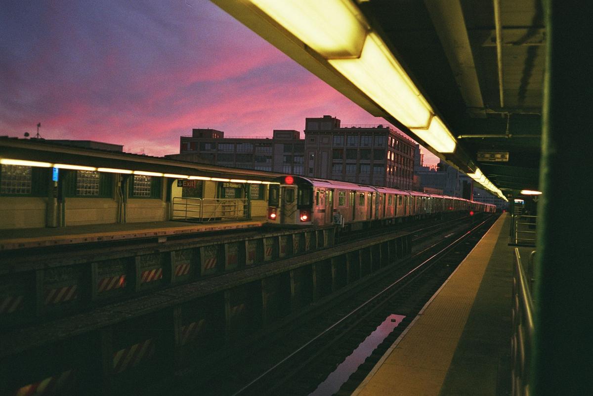 A subway train is stopped at a station platform under a vibrant pink and purple sky.