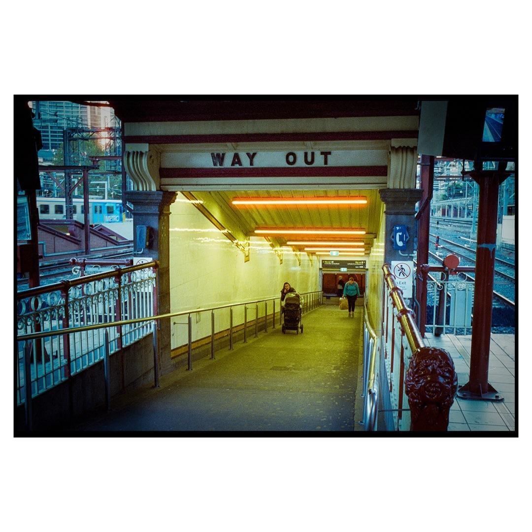 A woman pushes a stroller down a brightly lit underground walkway with a sign that reads "WAY OUT" above her.