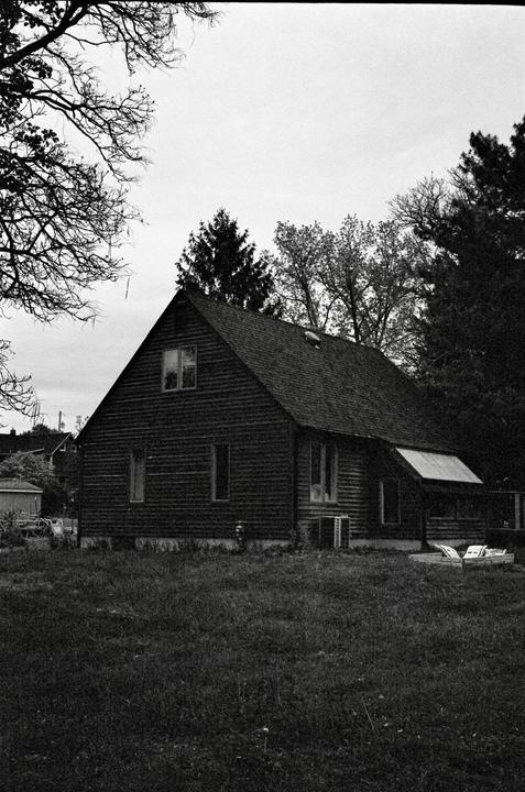 A dark wooden house with a steep roof stands in a grassy field surrounded by trees under a cloudy sky.