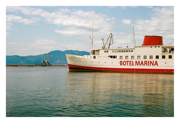 A white and red boat with "BOTEL MARINA" written on its side floats on water with mountains and cranes in the background.