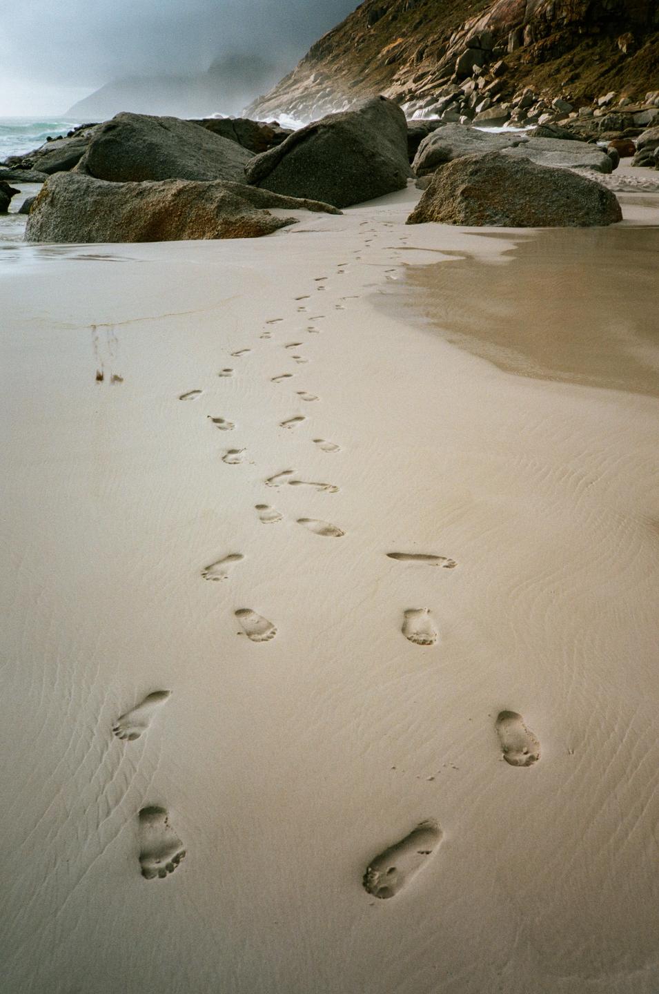 Footprints lead across a sandy beach towards large rocks and the ocean under a cloudy sky.
