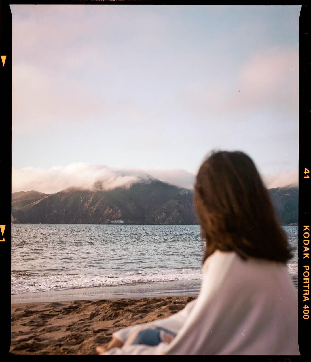 A woman sits on a sandy beach looking out at the ocean and a mountain range covered in clouds.