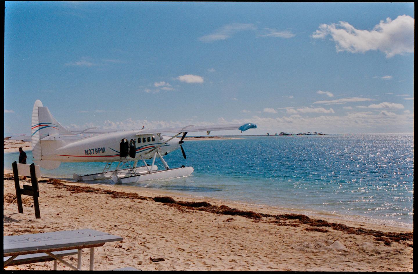 A white seaplane is parked on a sandy beach next to a clear blue ocean under a bright sky.