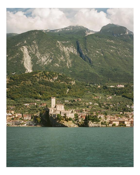 A castle stands on a rocky outcrop by a lake, with a town built into the hillside and mountains in the background.