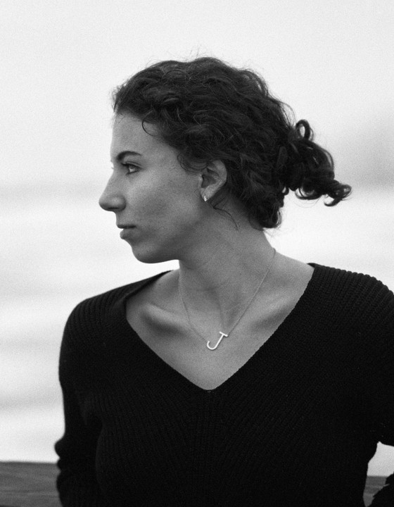 A woman with curly hair tied back wears a v-neck top and a necklace with a letter J pendant, looking to the left of the frame.