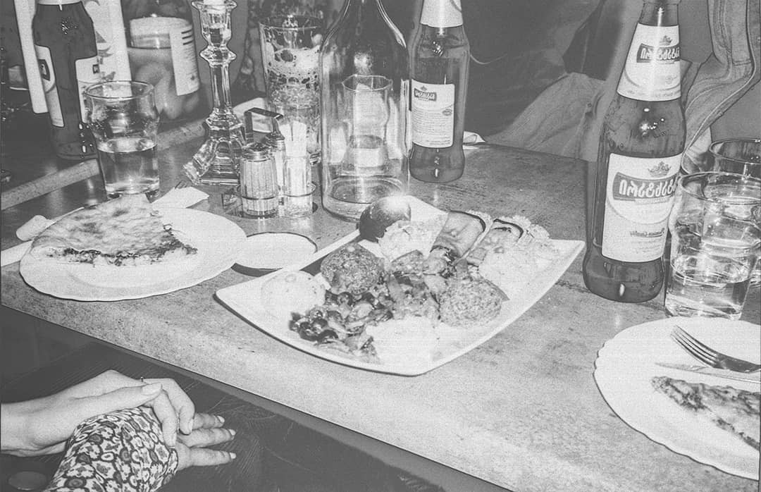 A table is laden with food, drinks, and a candelabra, with two hands clasped in the lower left corner.