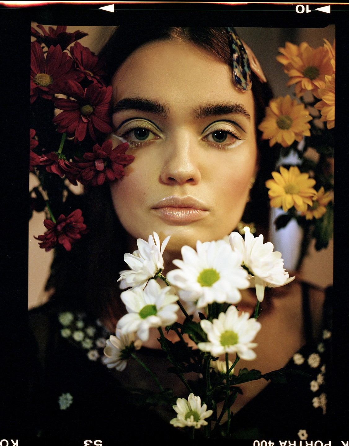 A woman with white eyeliner looks directly at the viewer, surrounded by red, yellow, and white flowers.