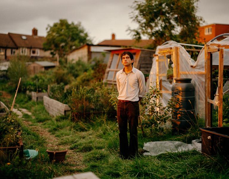 A man stands in a garden with greenhouses and houses in the background.