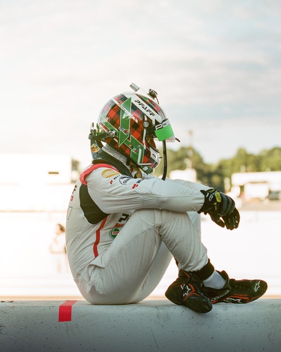A race car driver in a full suit and helmet sits on a concrete barrier.