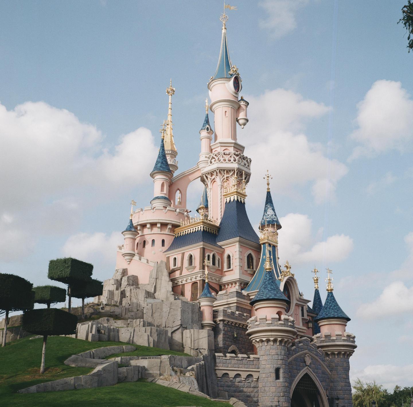 A pink and blue castle with multiple spires stands on a rocky hill with green grass and manicured trees under a cloudy sky.