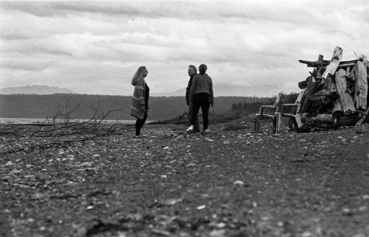 Three people stand on a rocky beach next to a large pile of driftwood and two wooden chairs.
