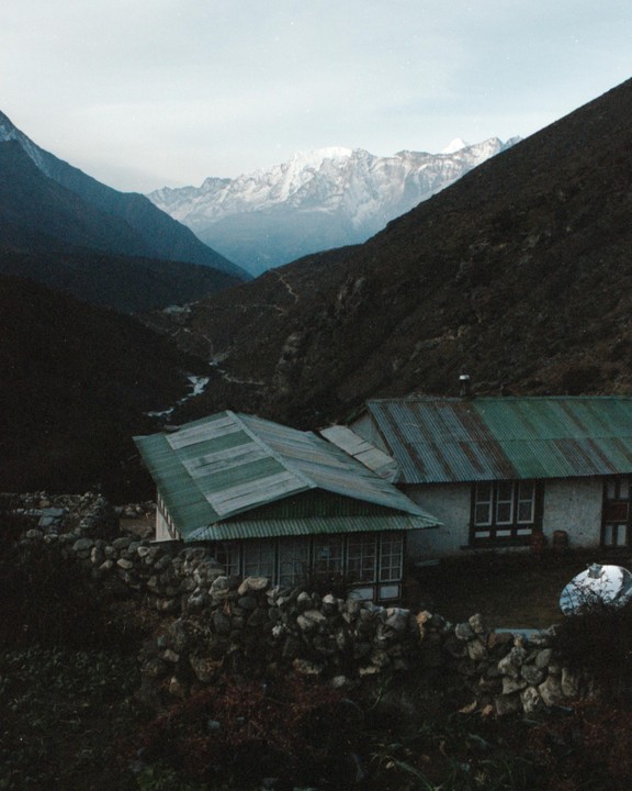 Two buildings with green roofs are nestled in a valley with snow-capped mountains in the distance.
