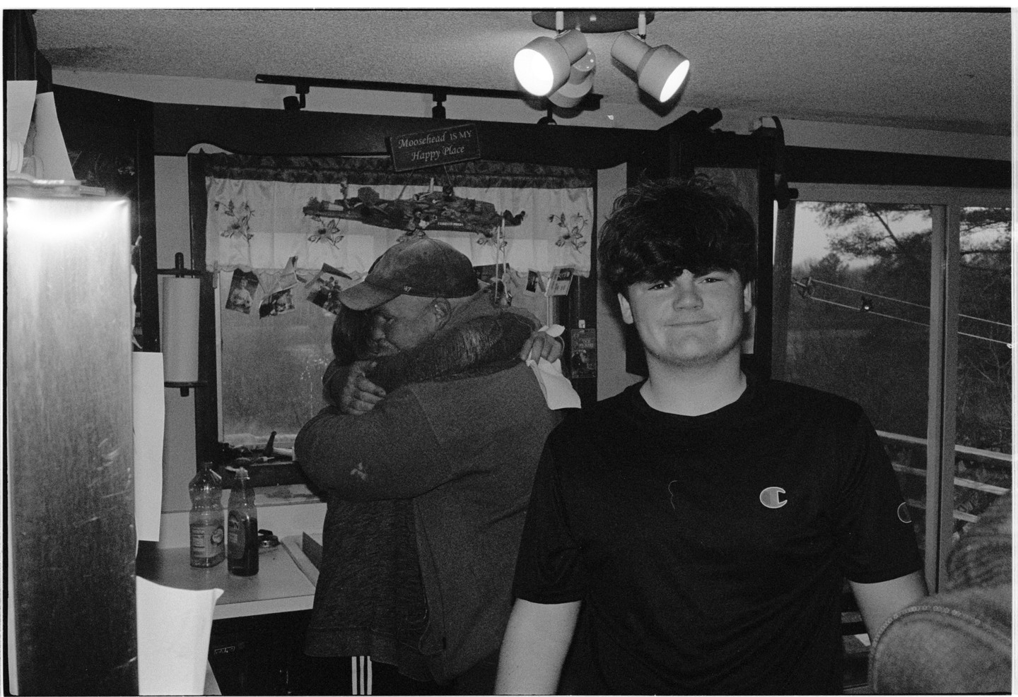 A man in a baseball cap hugs a woman while a young man smiles at the camera in a room with a window looking out to trees.
