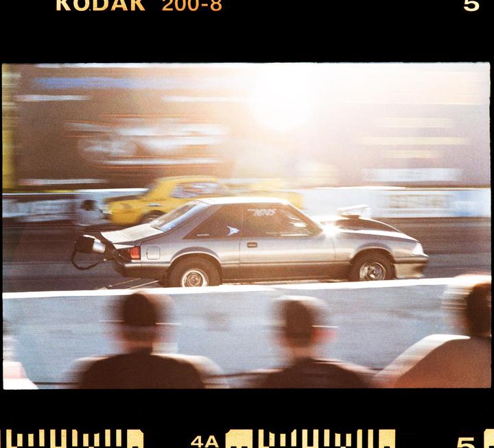 A silver race car speeds down a track with a yellow car in the background and spectators in the foreground.