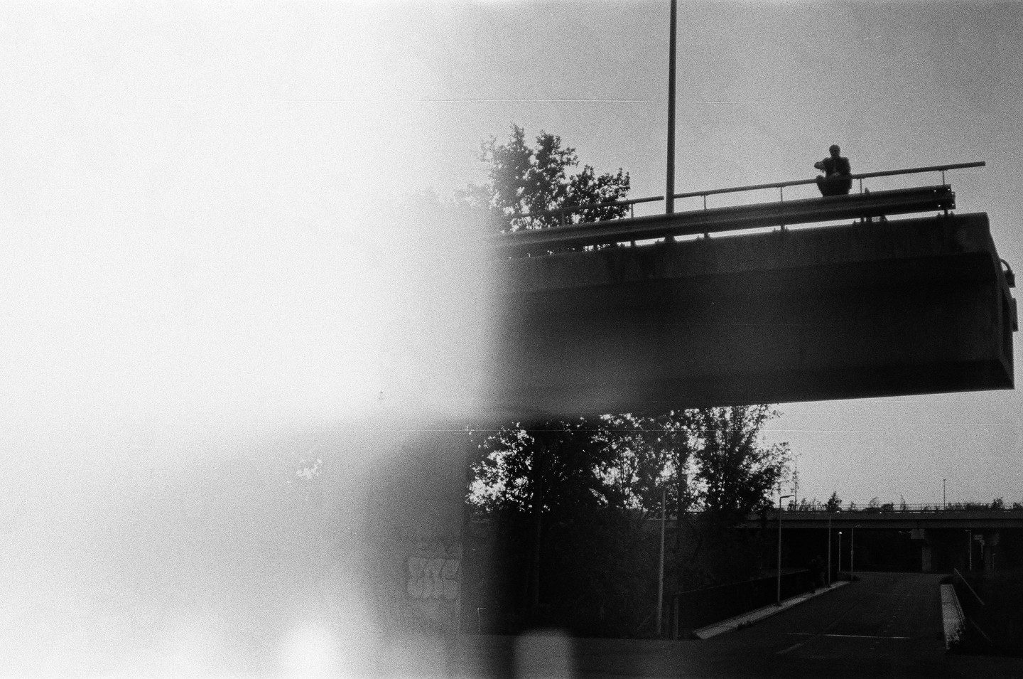 A man sits on a bridge railing above a road with trees and another bridge in the background.