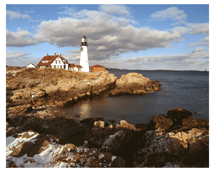 A white lighthouse and a house with a red roof stand on a rocky coast next to the ocean under a cloudy sky.