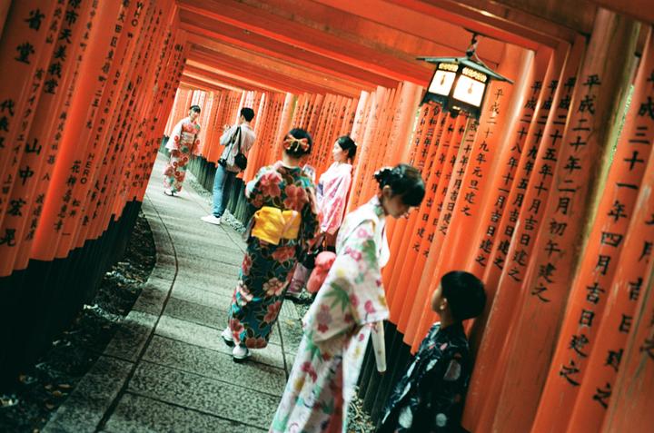 People in traditional Japanese clothing walk through a corridor of orange torii gates.