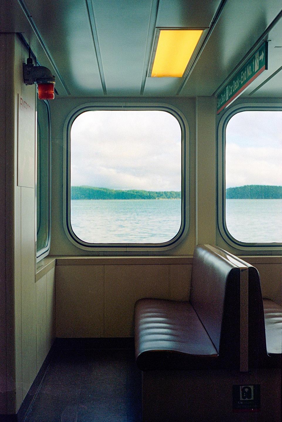 A ferry interior with a brown bench seat and windows looking out onto a body of water with a forested shore.