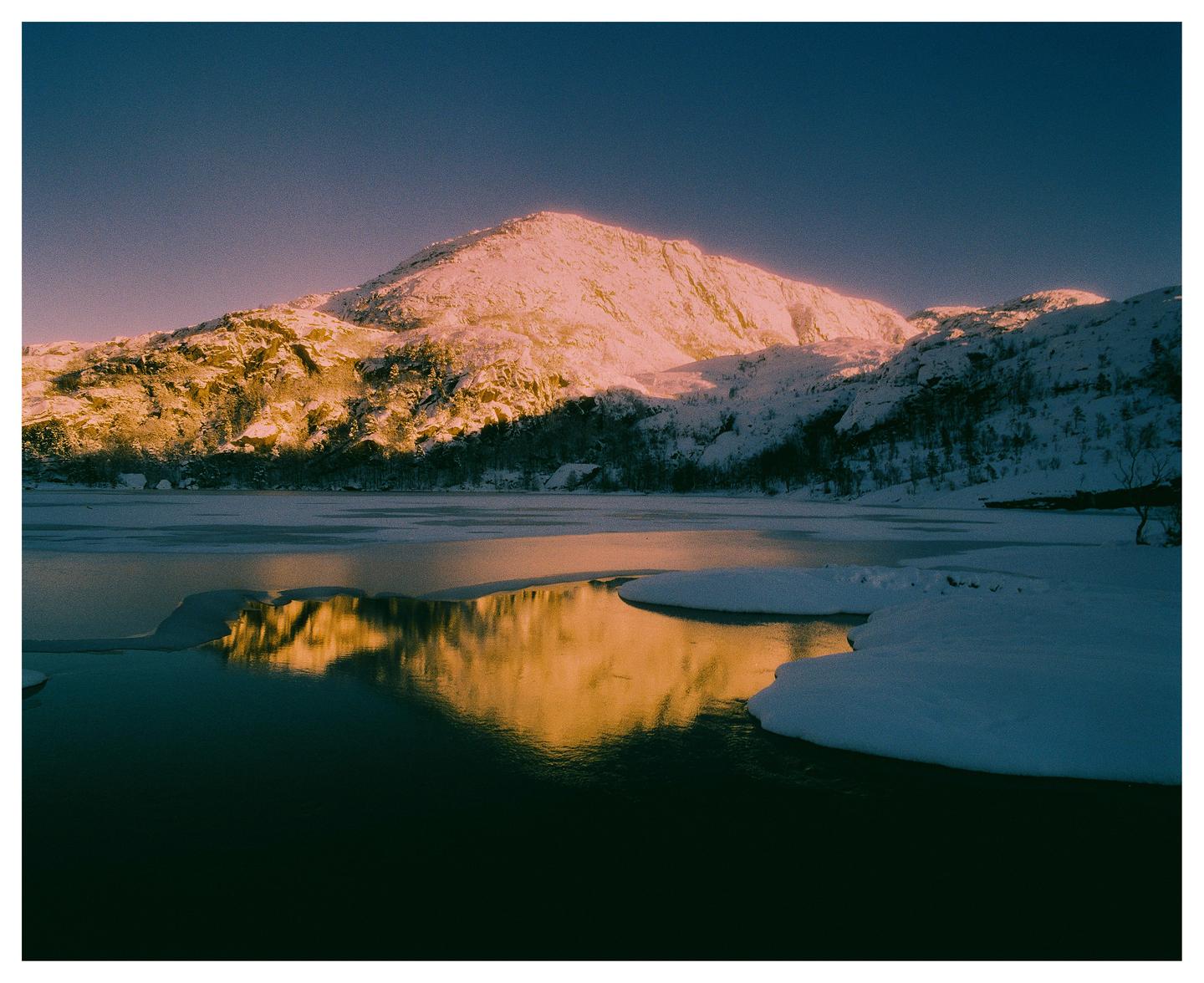 A snow-covered mountain reflects in a partially frozen lake under a clear sky.
