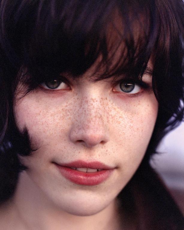 A woman with dark hair and freckles looks directly at the viewer.