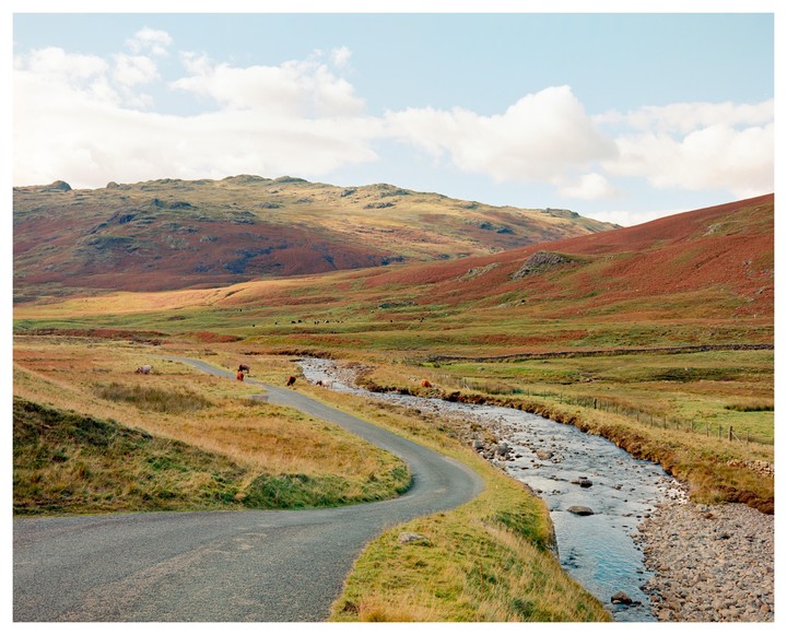 A winding road follows a river through a valley with hills covered in red and green foliage under a blue sky with clouds.