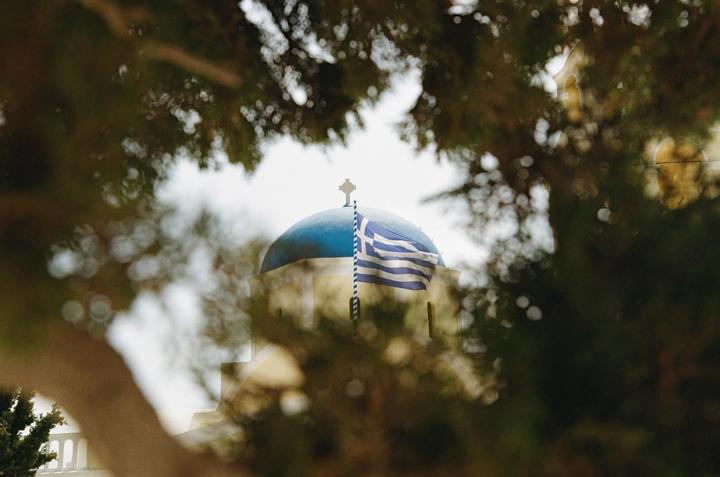 A Greek flag flies in front of a blue-domed building with a cross on top, framed by tree branches.