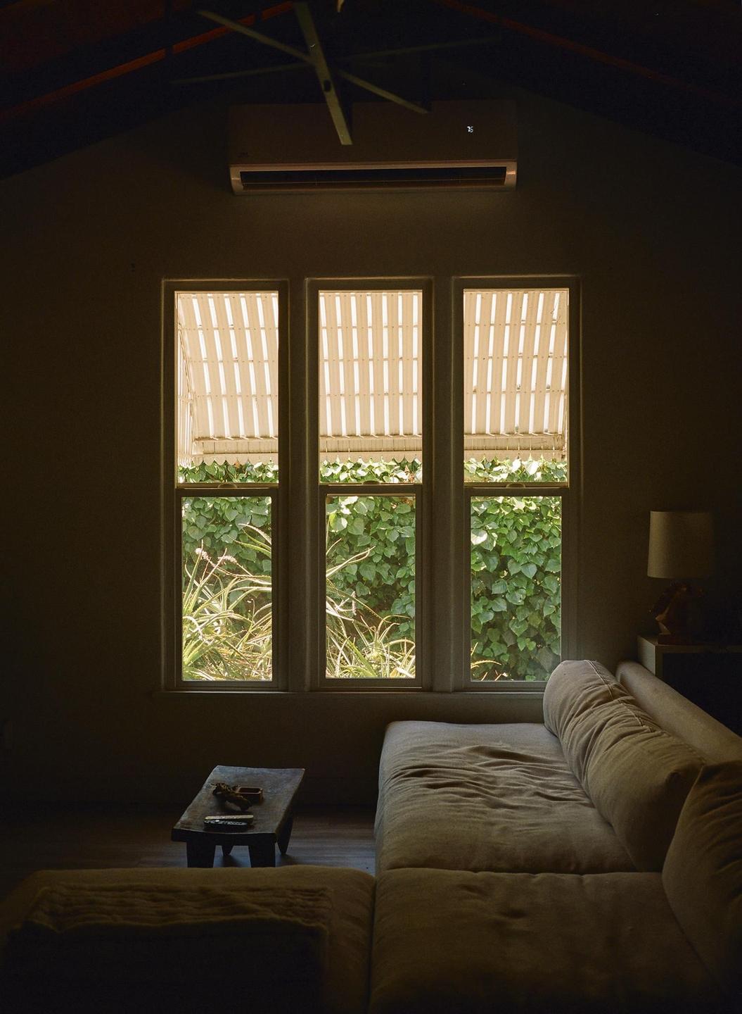 A dark living room with a large sofa and a small table is illuminated by three windows looking out onto green foliage.