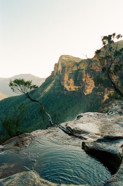 A small pool of water on a rocky cliff edge overlooks a vast valley with mountains and trees.