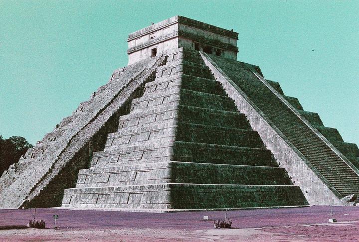 A large stone pyramid with multiple stepped levels and staircases rises under a clear sky.