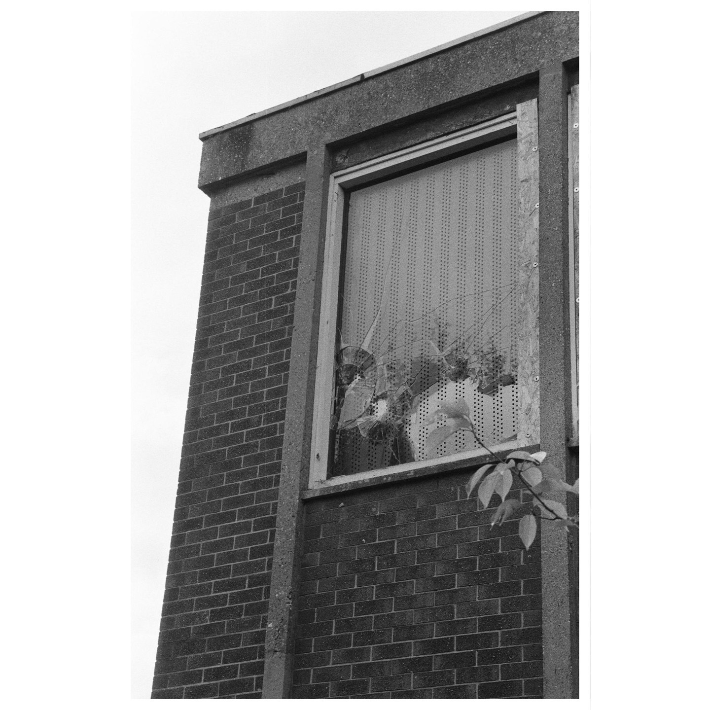 A broken window on a brick building is boarded up with a perforated panel.