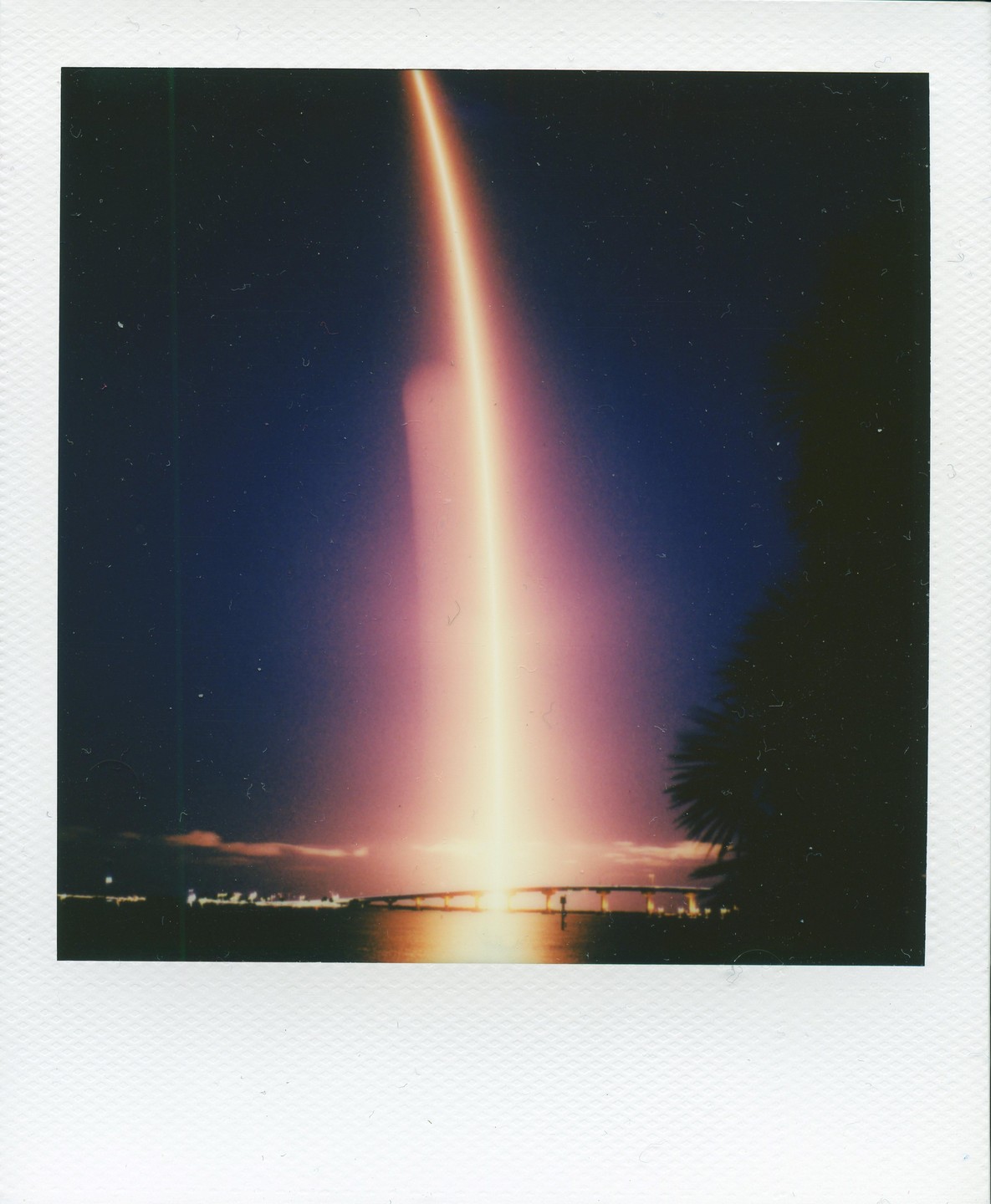 A long exposure captures the bright light trail of a rocket launch over a body of water with a bridge and city lights in the distance.