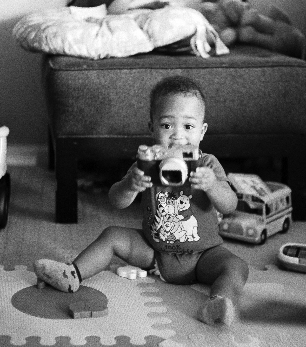 A young boy sits on a playmat, holding a toy camera and looking directly at the viewer.