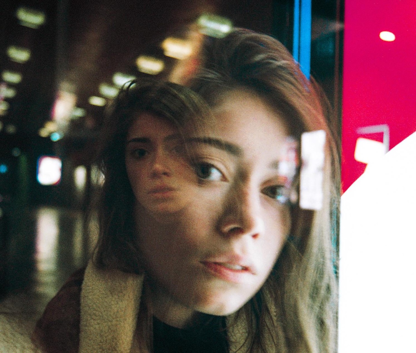 A woman's face is visible through a window, with another face reflected in the glass.