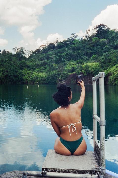 A woman in a bikini sits on a dock and takes a selfie with a phone, with a lake and forest in the background.