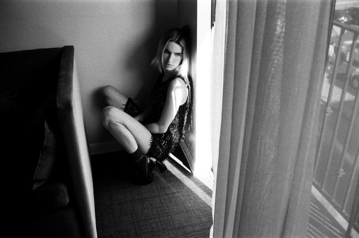 A woman crouches in a corner next to a window with sheer curtains and a balcony.
