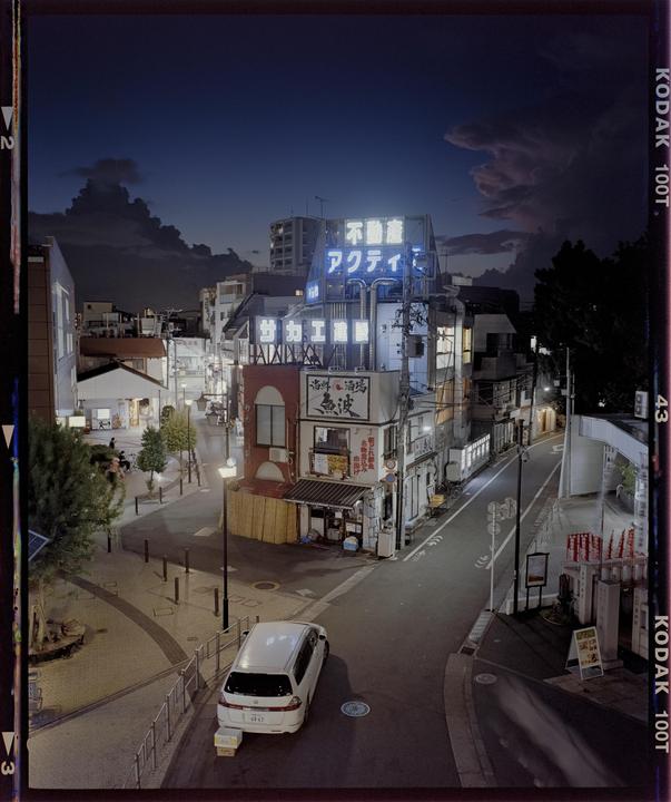 A street with buildings and a parked car is illuminated by neon signs and streetlights at dusk.