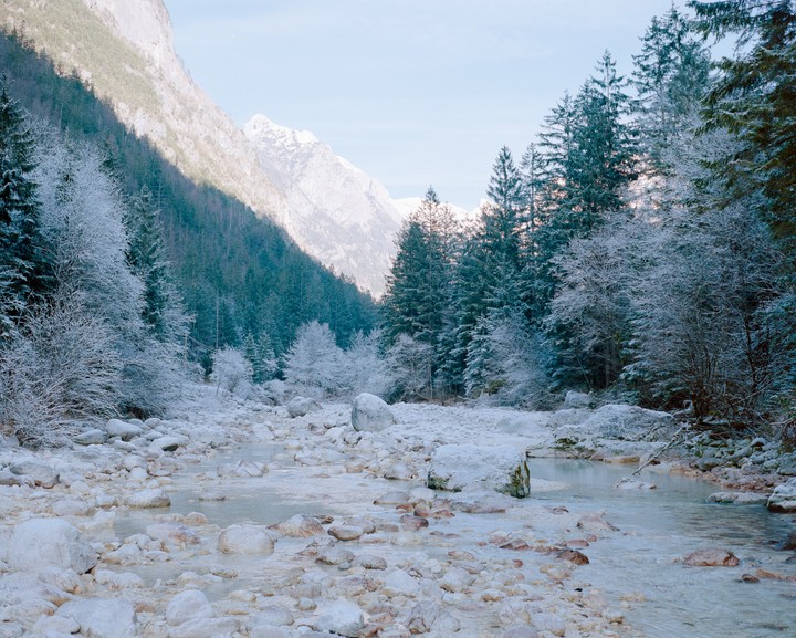 A frozen riverbed with snow-covered trees and mountains in the background.
