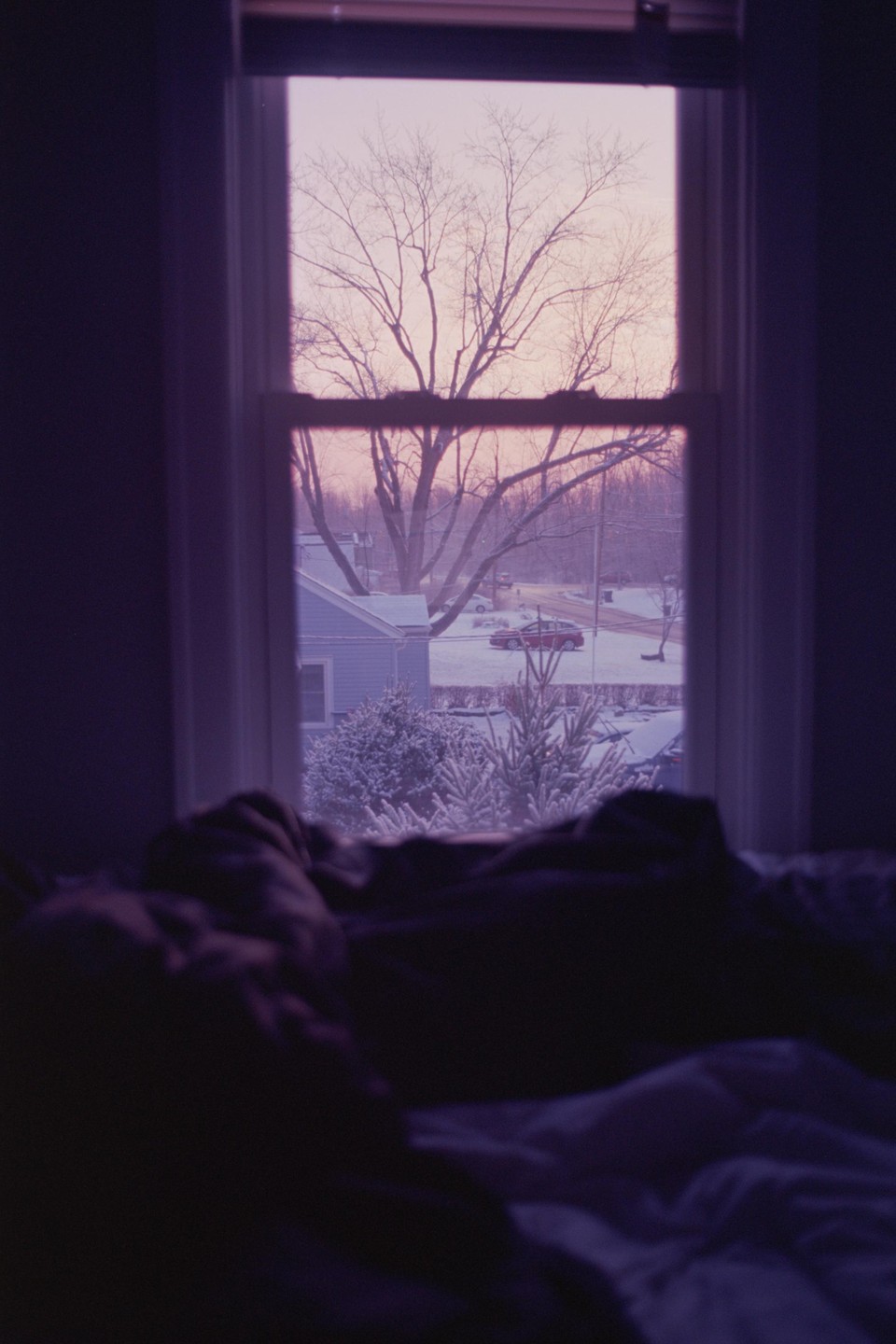 A bedroom window looks out onto a snowy street with a bare tree and a house.