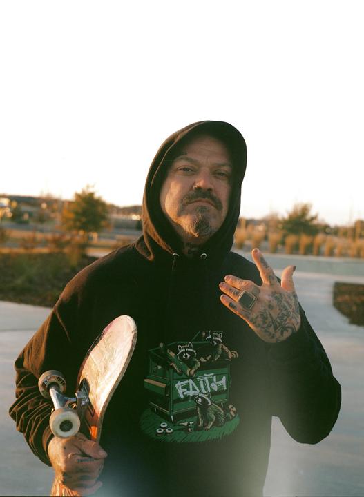 A man in a black hoodie with a skateboard under his arm poses for a portrait.