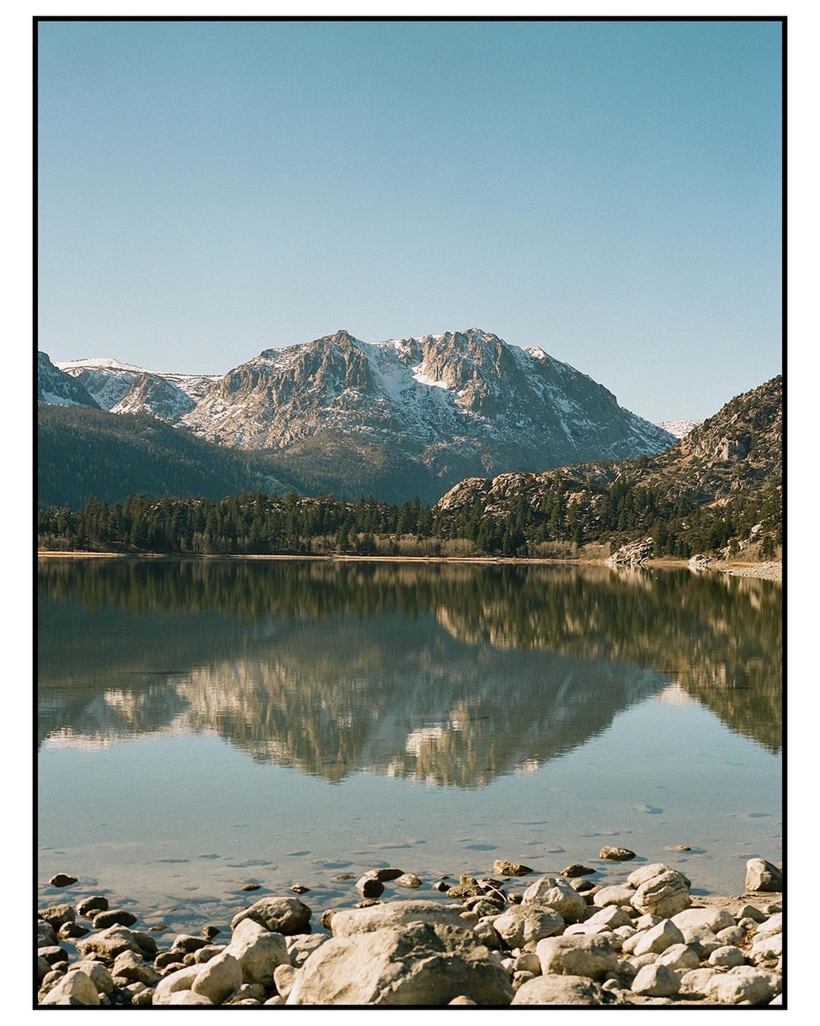 A snow-capped mountain range and a forest are reflected in a clear lake with a rocky shore.